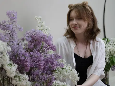 YukiPage smiles while wearing a white shirt and cat ears, posing next to large bunches of purple and white lilac flowers.