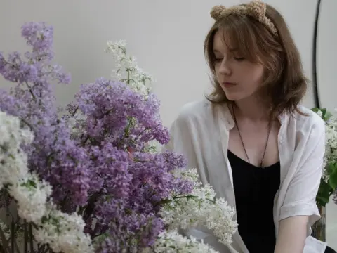 YukiPage wearing a white shirt and a fuzzy headband, sitting next to large bunches of purple and white lilacs.