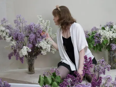 YukiPage sits on the floor among large bouquets of purple and white lilacs, wearing a black top and a white open shirt.