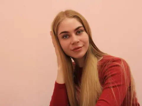 BarbaraNevill with long blonde hair wearing a red top, posing against a pink wall.