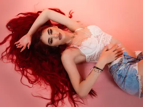 Woman with long wavy red hair wearing a white lace bralette and denim shorts lying on a pink background.