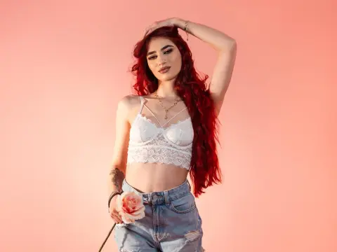 VixenRose — Red-haired woman in a white lace crop top and denim shorts posing against a pink background with a rose.