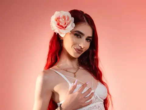 VixenRose — Beautiful woman with red hair and a pink rose accessory wearing a white lace top against a pink background.