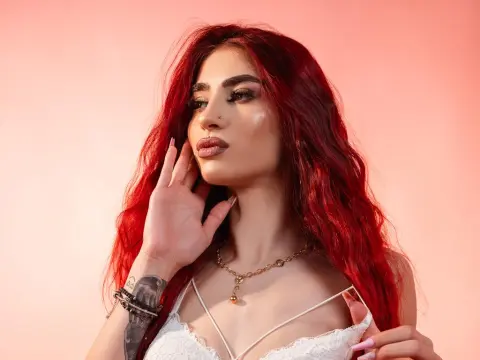 VixenRose — Glamorous woman with wavy red hair and gold necklace posing against a pink background.