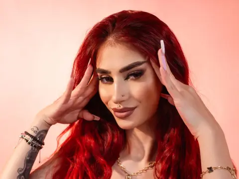 VixenRose — Glamorous woman with long red hair and bold makeup posing against a pink background.