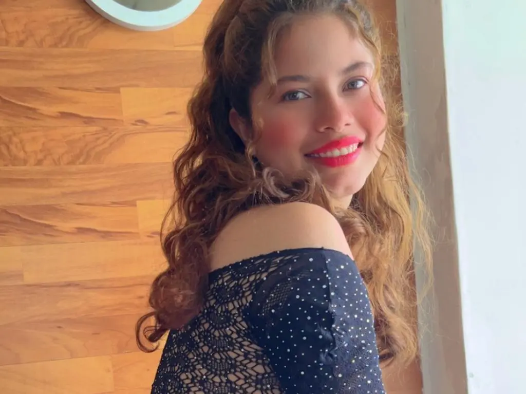EmilyCCm — Smiling young woman with wavy brown hair wearing a black lace polka dot top in a sunlit room.
