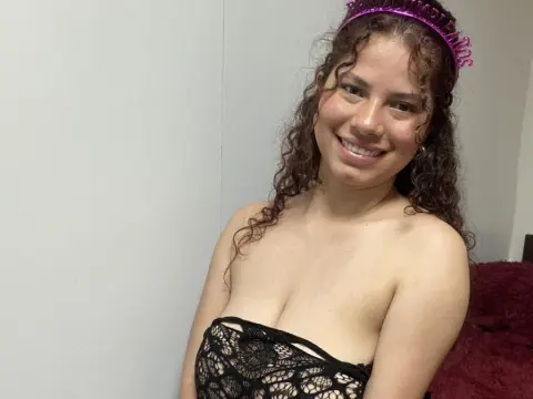 EmilyCCm — Smiling young woman with curly hair wearing a pink birthday tiara and black lace top in a bright room.