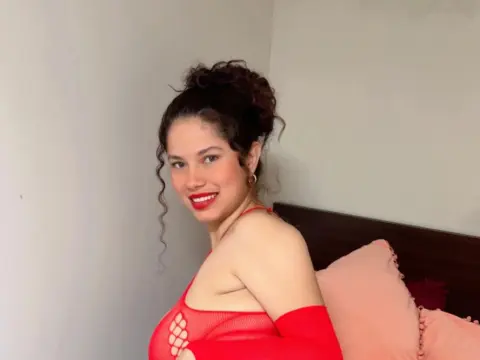 EmilyCCm — Smiling young woman in a red mesh outfit posing in a bedroom setting.