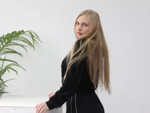 Blonde woman in a black velvet sweatshirt posing against a white wall with a green palm plant.