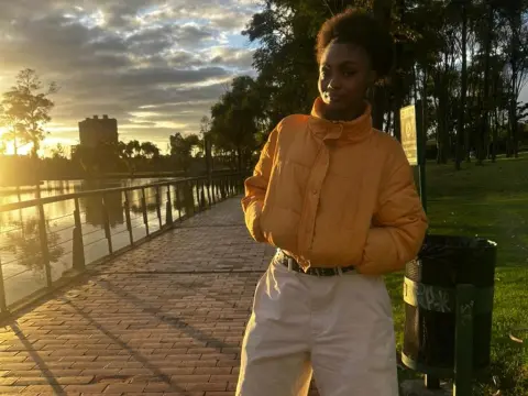 Asha_zuri69 poses outdoors in a bright orange puffer jacket and white pants during a sunset by a lake.