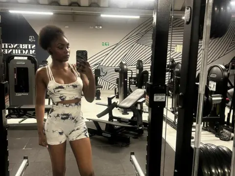 Asha_zuri69 poses in a gym wearing a white tie-dye sports bra and matching shorts, showing her toned stomach and hair in an updo.