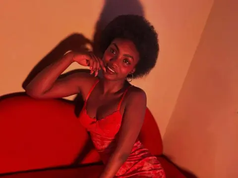 Asha_zuri69 poses on a red couch wearing a red lace camisole, showing her dark hair and hoop earrings against a warm background.