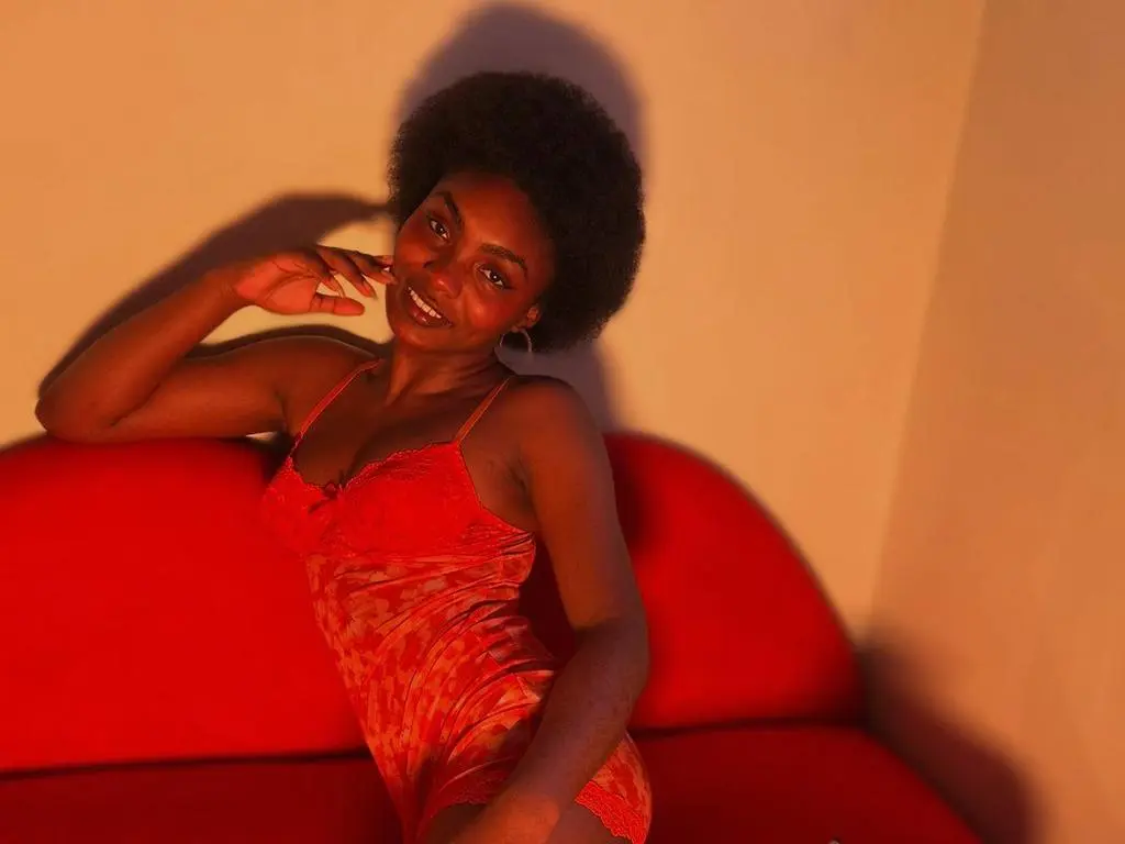 Asha_zuri69 — Asha_zuri69 wearing a red lace teddy, sitting on a red sofa with her hand near her face and her hair in a large afro.