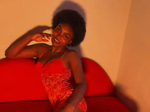 Asha_zuri69 wearing a red lace teddy, sitting on a red sofa with her hand near her face and her hair in a large afro.