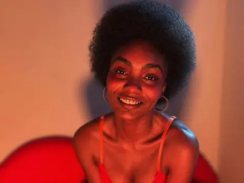 Asha_zuri69 smiles while wearing a red camisole and large hoop earrings, showing off her short afro hair against a plain wall.