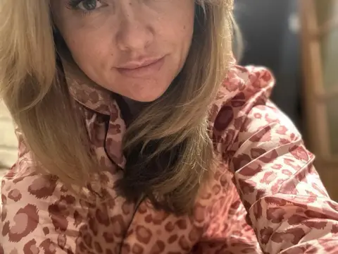 Blonde woman in pink leopard print silk pajamas smiling in a cozy indoor setting.
