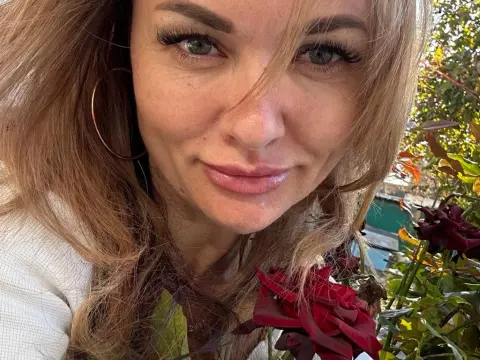 Smiling blonde woman with gold hoop earrings posing near red roses in a sunny garden.