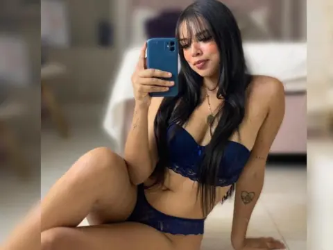 Young woman in navy blue lace lingerie sitting in a bright bedroom taking a mirror selfie.