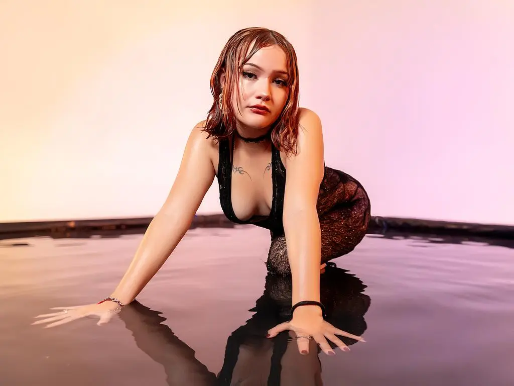 Abril_Cardenas poses in a black halter top and sparkly skirt while kneeling in shallow water against a pink background.