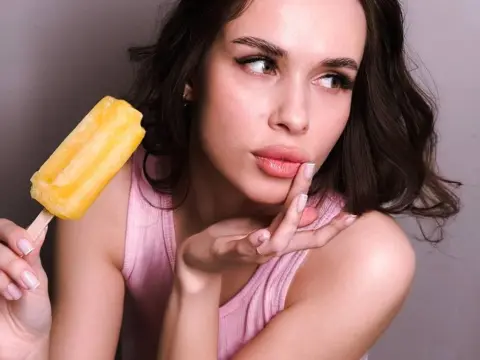 Young woman in a pink tank top enjoying a refreshing mango popsicle against a neutral studio background.