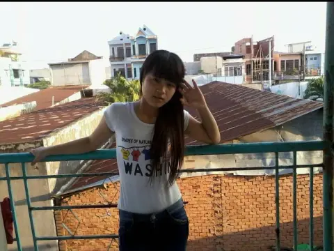BabySweet18x — Young woman in a white graphic t-shirt and jeans leaning on a balcony railing overlooking a city rooftop.