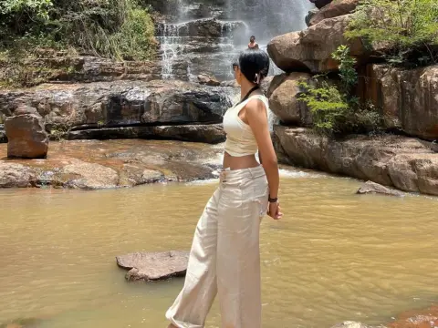 BiancaDream — Woman in white linen outfit standing by a rocky waterfall in a serene natural jungle setting.