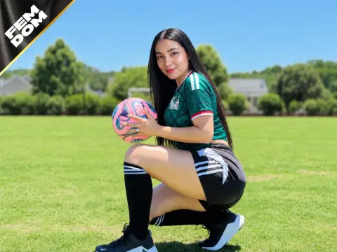 PoppyKensingtonOG squats on a green grass field while holding a pink and blue soccer ball, wearing a green jersey and black athletic shorts.