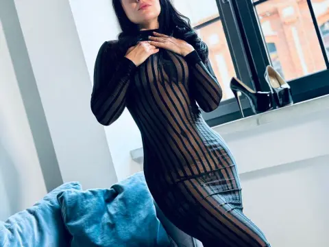 Elegant woman in a sheer black striped maxi dress posing by a bright window in a modern bedroom.