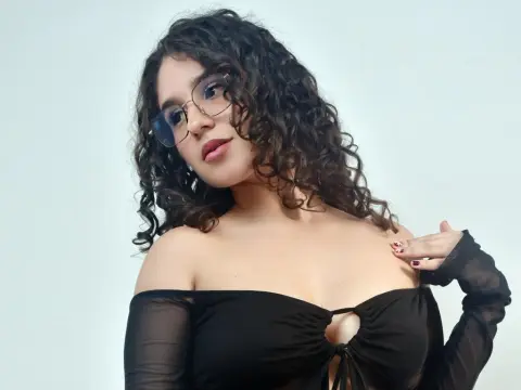 Young woman with curly hair and glasses wearing a black off-the-shoulder top in a bright studio setting.