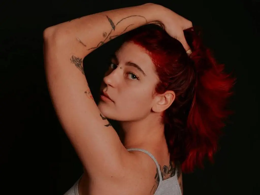 Red-haired woman with arm tattoos posing in a grey tank top against a dark background.