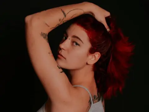Red-haired woman with arm tattoos posing in a grey tank top against a dark background.