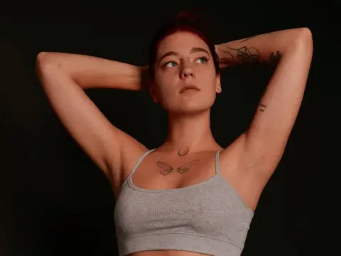 Young woman with red hair and chest tattoos wearing a grey ribbed crop top against a dark studio background.