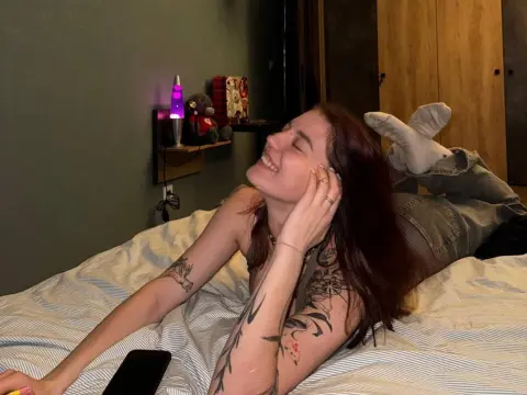 Smiling woman with red hair and tattoos relaxing on a bed in a cozy, dimly lit bedroom.