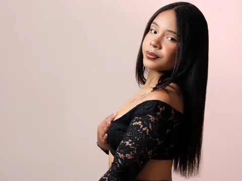 Young woman with long dark hair wearing a black lace off-the-shoulder top against a soft pink background.