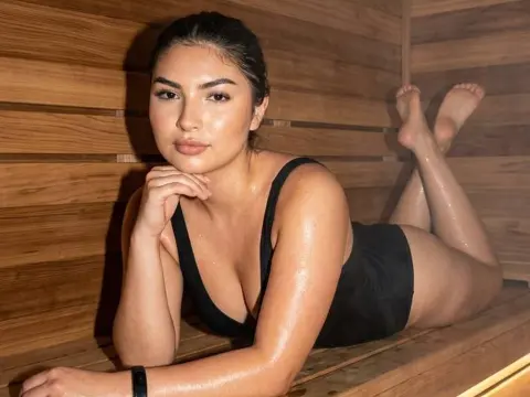 AzzaLeeX lies on a wooden sauna bench wearing a black bodysuit, showing off her wet, glowing skin and dark hair.