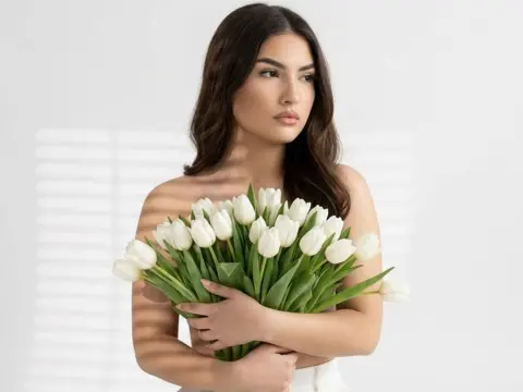 AzzaLeeX holds a large bouquet of white tulips against her bare chest, looking off to the side with her long dark hair down.