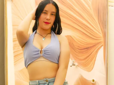 AlairaAura poses in a light blue halter top and denim jeans, running her hand through her long black hair.