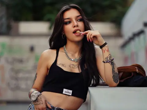 VanesaHaze licks a lollipop while wearing a black crop top, showing her long dark hair, tattoos, and silver chain necklace.