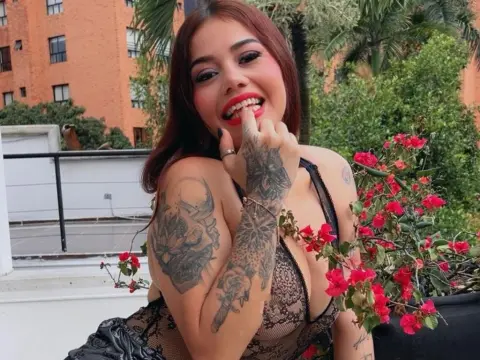 DarkHoneeey smiles while biting her finger, showing off her tattoos and black lace bodysuit in a garden setting.