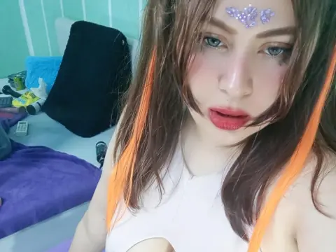 Close-up selfie of a woman with long brown and orange hair, glitter makeup, and a white top in a bedroom setting.