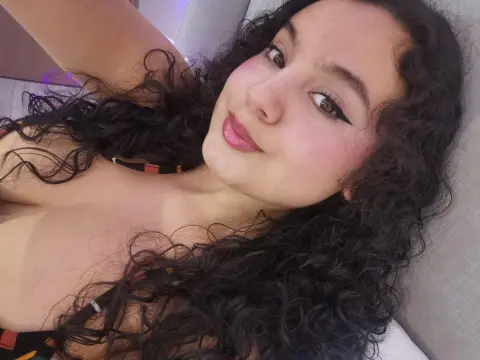 Smiling woman with curly dark hair and winged eyeliner wearing a black patterned top in a cozy indoor setting.