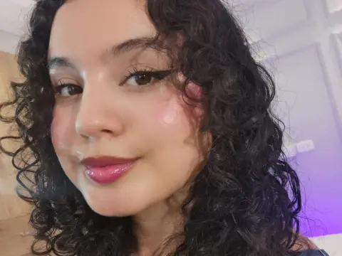 Close-up portrait of a smiling woman with curly dark hair and winged eyeliner in soft indoor lighting.