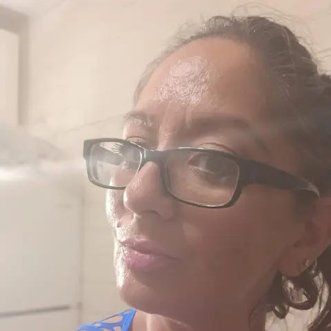 Close-up portrait of a smiling woman with glasses in a bright, sunlit indoor setting.