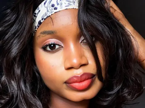 GailRose poses with her dark wavy hair held back by a white patterned headband, showing her face and red lipstick.