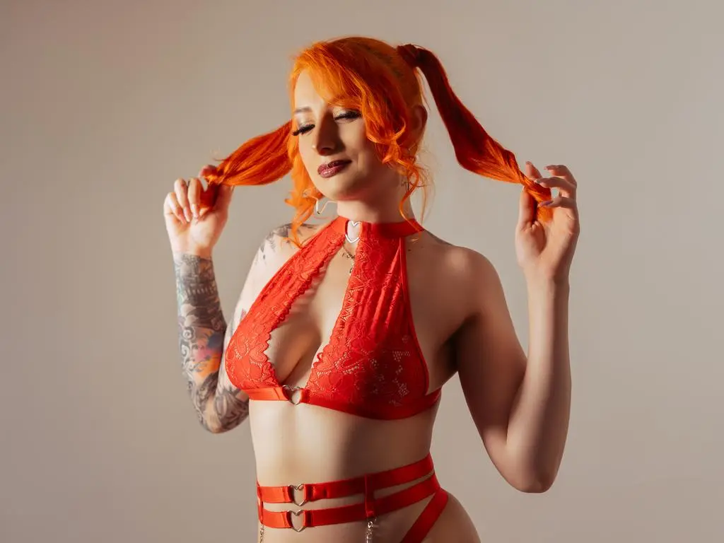VenusMontiel, wearing a red lace halter bikini and red garter straps, poses with her orange hair in pigtails against a plain background.