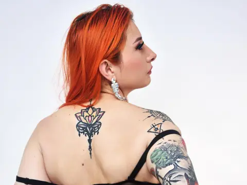 VenusMontiel shows her bright orange hair and back tattoos while posing in a black strap top against a white background.
