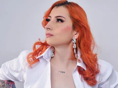 VenusMontiel, a woman with bright orange hair and long silver earrings, wearing an open white button-down shirt against a plain white background.