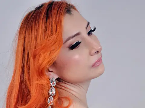 VenusMontiel poses with her bright orange hair and long eyelashes, wearing large crystal drop earrings against a plain white background.