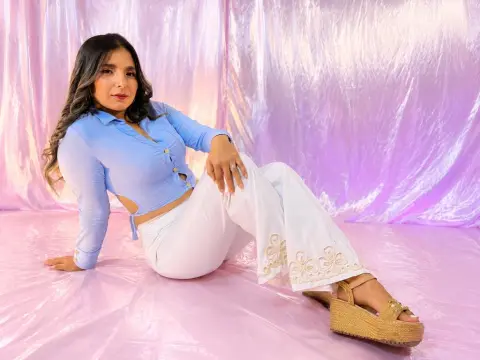 Young woman in a light blue button-down shirt and white flared trousers posing against a pink shimmering backdrop.