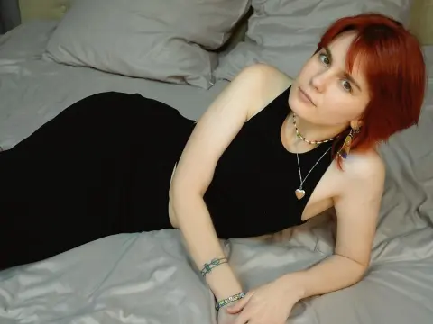 AvroraVance18 wearing a black tank top, lying on a grey bed with her red hair down and wearing a heart necklace.